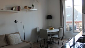A modern living room with a sofa, a dining table with chairs, a lamp, and a window with a view. - Charming two-room refurbished 50m from the sea (Houlgate)
