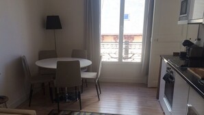 A small, open-plan living and dining area with a kitchen, a dining table with chairs, a patterned rug, and a view through a window. - Charming two-room refurbished 50m from the sea (Houlgate)
