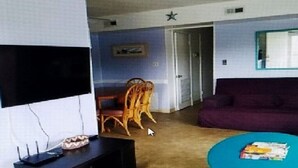 Una sala de estar con televisor de pantalla plana, mueble negro para entretenimiento, sofá morado, mesa de centro verde azulado y una silla de madera.