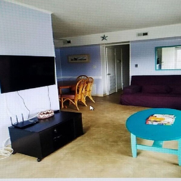 A living room with a flat-screen TV, a black entertainment unit, a purple sofa, a teal coffee table, and a wooden chair.