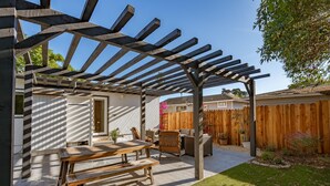 Eine überdachte Terrasse mit einem Holztisch und Stühlen, einer Pergola darüber und einem von einem Zaun umgebenen Garten.