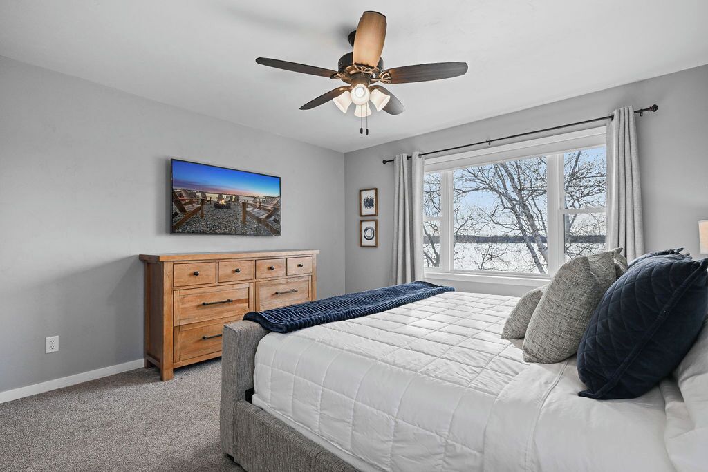 A bedroom with a bed, a ceiling fan, a dresser, a window with curtains, and a painting on the wall.