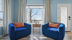 Una habitación con dos sofás azules, una pequeña mesa de madera y vistas al exterior de un árbol y agua.