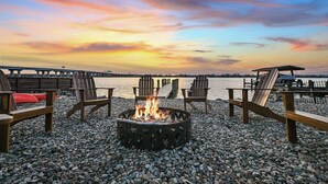 Un hogar con fuego rodeado de sillas Adirondack en una playa de guijarros, con vista a un puente y al atardecer.