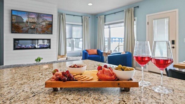 Una zona de comedor con una tabla de madera que presenta diversos quesos, carnes y frutas, junto con dos copas de vino tinto.