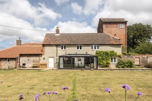 A two-story house with a conservatory, a stone wall, and a grassy yard with purple flowers.