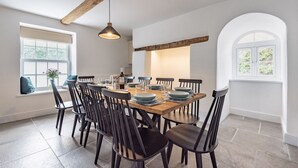 Un coin repas avec une table en bois dressée pour six personnes, entourée de chaises sombres. Un luminaire suspendu se trouve au-dessus de la table, et la pièce est dotée de murs blancs et d’un plafond avec une poutre en bois.