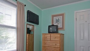Une chambre à coucher avec un lit, une commode en bois, un téléviseur posé dessus, un tableau encadré et une porte.