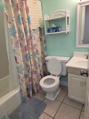 A bathroom with a toilet, a bathtub, a shower curtain with a shell pattern, a white sink, and a small shelf with toiletries.