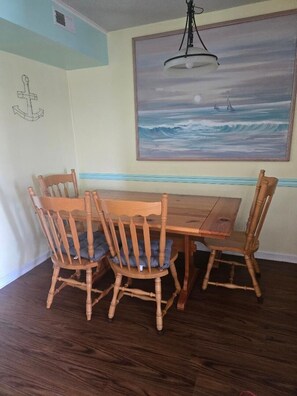 A dining area with a wooden table and chairs, a painting of a seascape on the wall, and a ceiling light fixture.