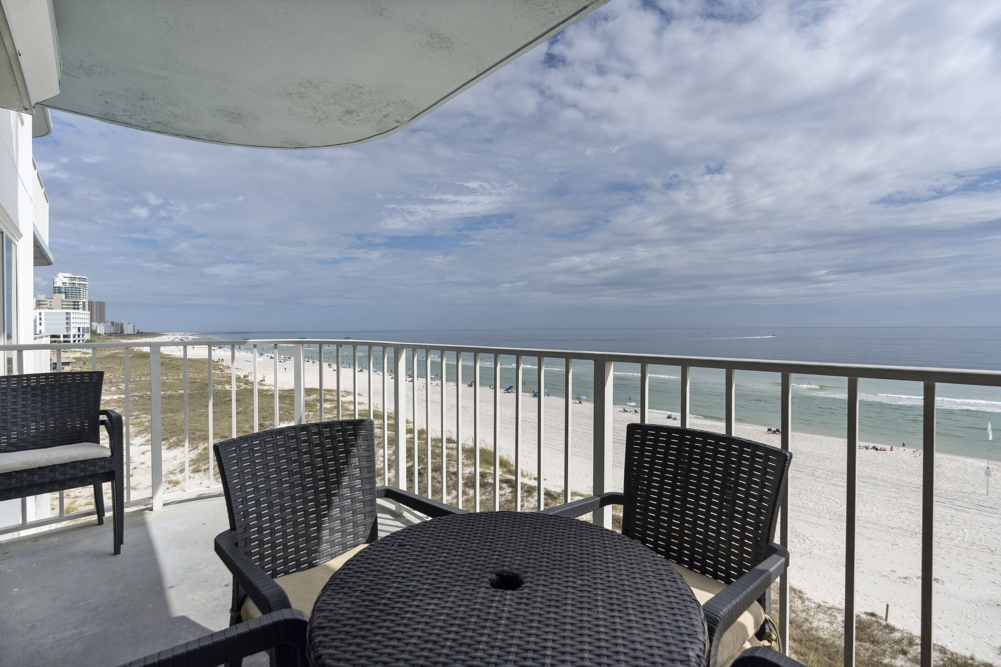 Een balkon met rieten stoelen en een tafel, met uitzicht op een strand met een hek en zee.