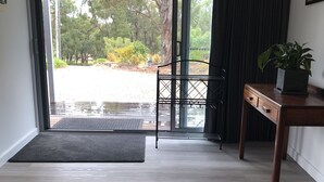 A room with a wooden floor, a sliding glass door leading to a garden, a black metal chair, a wooden table with a plant, and a dark curtain.