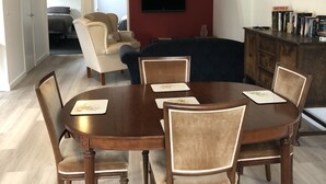 A modern living room with a round wooden dining table, cushioned chairs, a dark sofa, and a mounted flat-screen TV.
