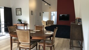 A modern living room with a dining area, featuring a wooden dining table and chairs, a sofa, a TV, and a bookshelf.