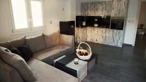 Un salon avec un canapé beige, une table basse et un panier de fruits. Il y a également un coin cuisine avec un plan de travail en bois et un four encastré.