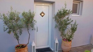 Une porte blanche ornée d’une fenêtre en forme de losange, flanquée de deux oliviers en pot et de deux lanternes blanches.