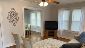 A living room with a ceiling fan, a TV on a wooden stand, a beige sofa, and a dining area with chairs and a table. - Home Town Getaway - 2 bed 1 bath home - close to town! (Paris)