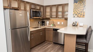 A modern kitchen with stainless steel appliances, wooden cabinets, a microwave, and a dining area with a round table and chairs. - Accessible Studio Suite, 1 King with Tub (Humble)