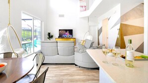 A modern living room with a sofa, a dining area with a table and chairs, a kitchen counter with food and drinks, and a staircase leading to an upper level. - SALT WATER POOL*EXPANSIVE POOL DECK* LED POOL FOUNTAIN JETS (kissimmee)