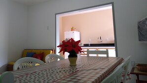 A dining area with a table set for two, a vase with red flowers, and a view of a kitchen through an open doorway. - Apartment 200 meters from the beach in Imbituba, SC. (Imbituba)