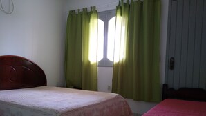 A bedroom with two beds, a window with green curtains, and a wooden headboard. - Apartment 200 meters from the beach in Imbituba, SC. (Imbituba)
