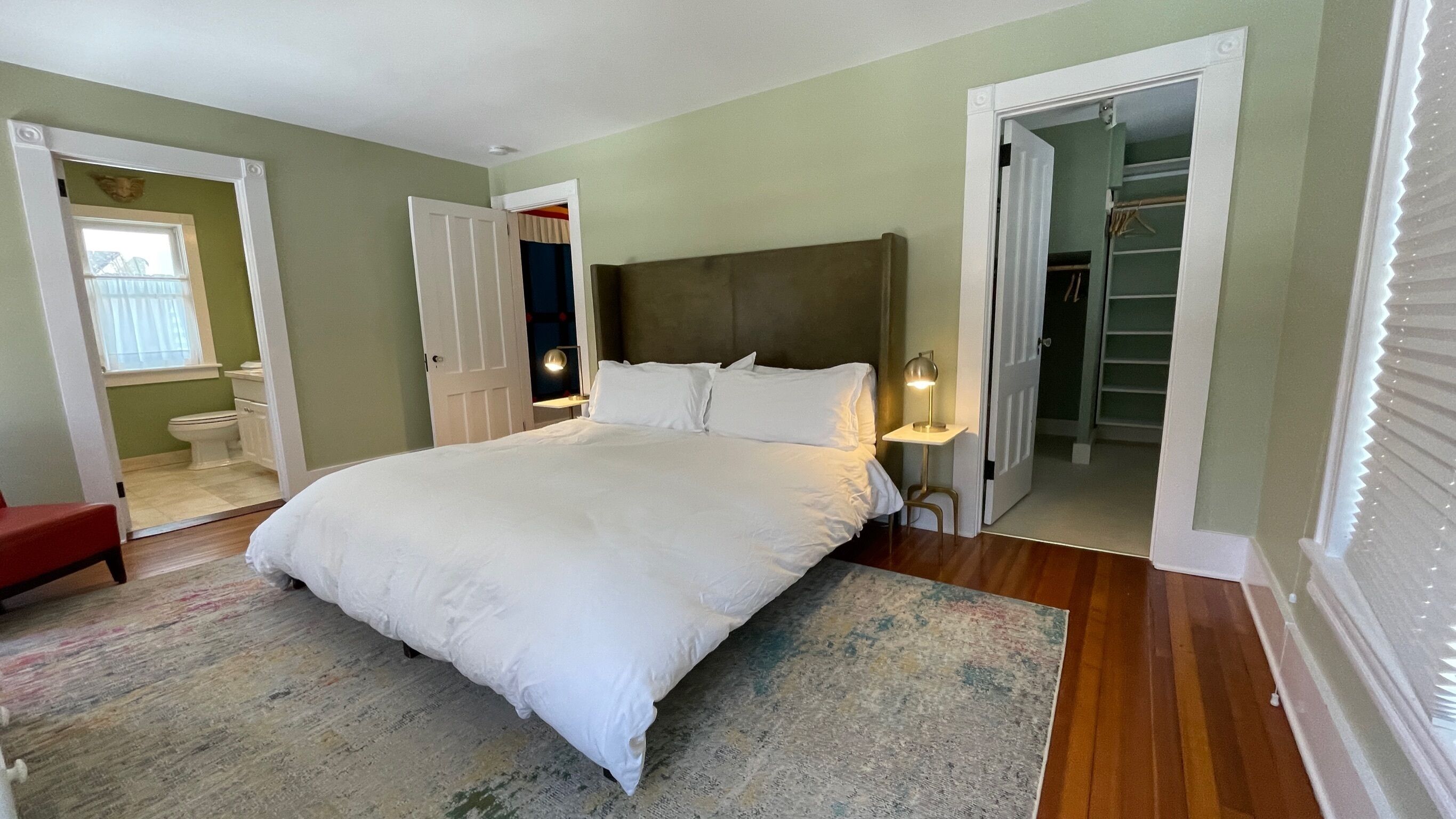 A bedroom with a large bed, a bedside table with a lamp, a red chair, and a view of a bathroom and closet.