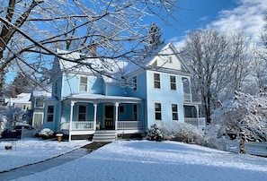 A two-story blue house with a porch, surrounded by snow-covered trees and a clear sky.