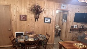 Una habitación tipo cabaña con interior de madera, una mesa de comedor con sillas, un televisor montado en la pared y un letrero que indica cabaña acogedora.