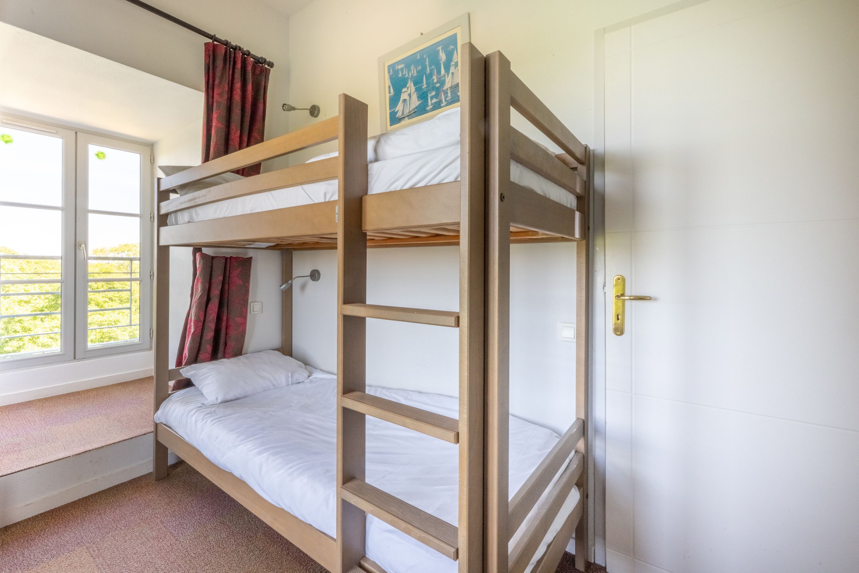 Une chambre avec un lit superposé, une fenêtre, une porte et un tableau encadré au mur.