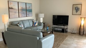 A living room with a grey sofa, a flat-screen TV, and a coffee table. There are two lamps, a starfish decoration, and a framed picture on the wall.