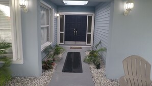 A house entrance with a dark front door, white sidings, and a black mat leading to the door. There are two wall-mounted lights and a chair to the right.