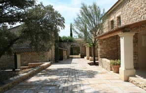 Ein Steinweg führt durch einen Innenhof mit traditionellen Steinbauten, Topfpflanzen und Bäumen.