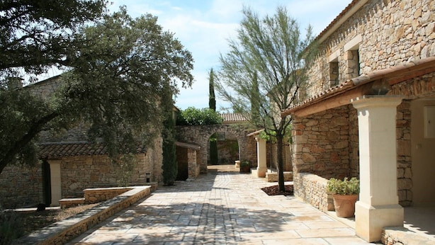 Un sendero de piedra atraviesa un patio con edificios de piedra tradicionales, plantas en macetas y árboles.