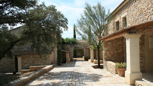 A stone pathway leading through a courtyard with traditional stone buildings, potted plants, and trees. - Appart Duplex en Provence | Piscine Saisonnière (Gordes)
