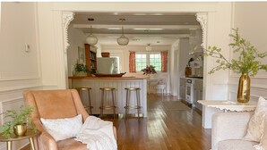 Un salón con un sillón de cuero marrón, un sofá blanco y una mesa de centro de madera con libros. Se observa una cocina al fondo con un refrigerador, estufa y taburetes de bar.