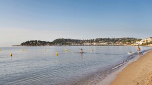 A beach with calm waters, a few people, and a distant hill with buildings. - Studio Climatisé | 10 Minutes de la Plage! (Cavalaire-sur-Mer)