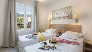 A hotel room with two beds, a large window with curtains, a painting on the wall, and a nightstand with a lamp. - Studio Climatisé | 10 Minutes de la Plage! (Cavalaire-sur-Mer)