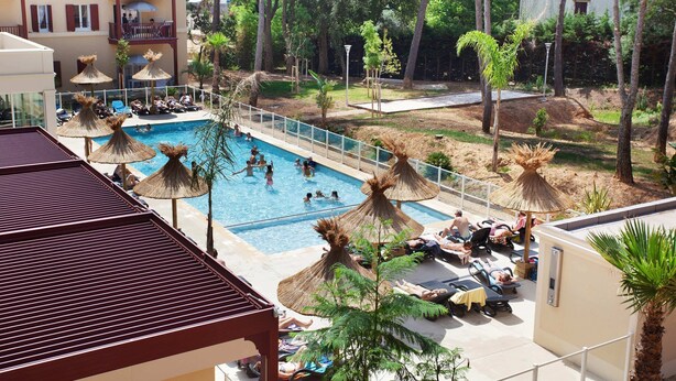 Un área de piscina con sillones de descanso y sombrillas, rodeada de árboles y un edificio con balcones.