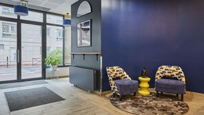 Un espace d’accueil moderne avec un mur d’accent bleu, deux fauteuils à motifs, une petite table d’appoint jaune et une grande fenêtre donnant sur la rue.