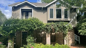 A two-story house with a brick foundation, a front porch, and a garden with various plants and flowers.