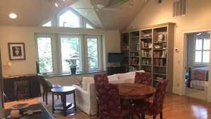 A cozy living room with a dining area, large windows, a bookshelf, and a ceiling fan.