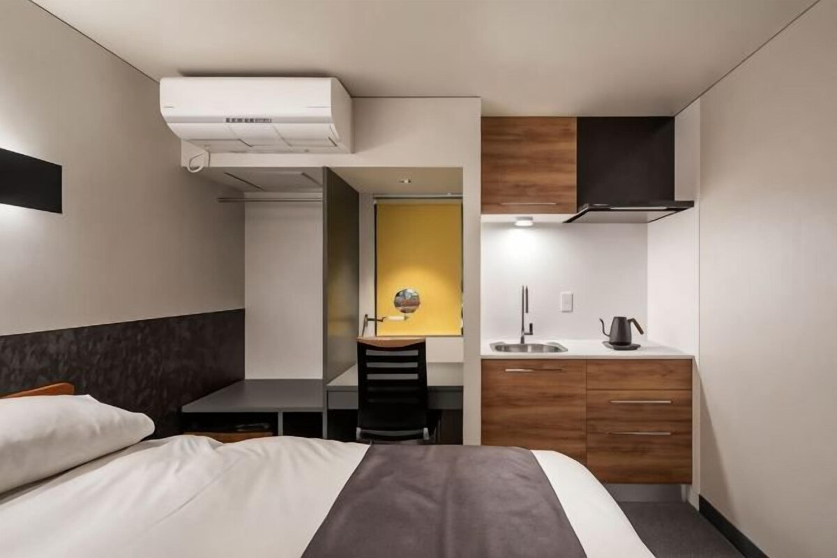 A hotel room with a bed, a desk, a chair, a small kitchenette, and a bathroom.