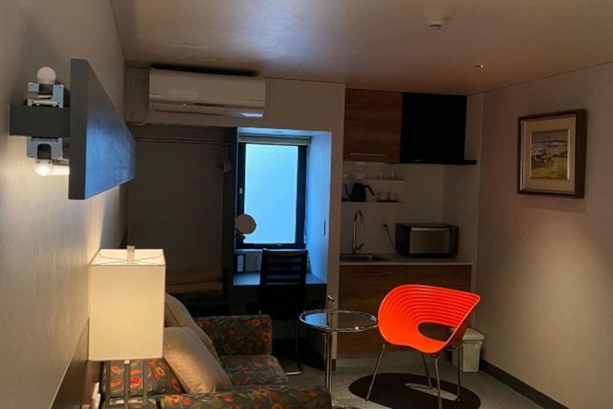 A compact hotel room with a sofa, a small table, a red chair, a TV, and a kitchen area visible through an open door.