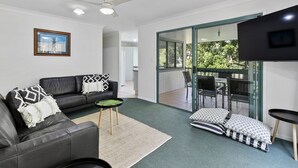 En moderne stue med en sort lædersofa, et lille rundt bord og et grønt tæppe. Der er en skydedør, som fører ud til en udendørs terrasse med et bord og stole.