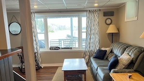A cozy living room with a wooden coffee table, a sofa with cushions, a lamp, and a view of the outdoors through a window. - Relaxing Lake House - Enjoy the Sun and Fun!!! (Erieville)
