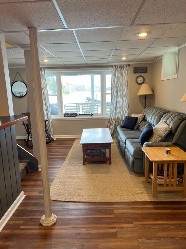 A cozy living room with a wooden coffee table, a sofa with cushions, a lamp, and a view of the outdoors through a window. - Relaxing Lake House - Enjoy the Sun and Fun!!! (Erieville)