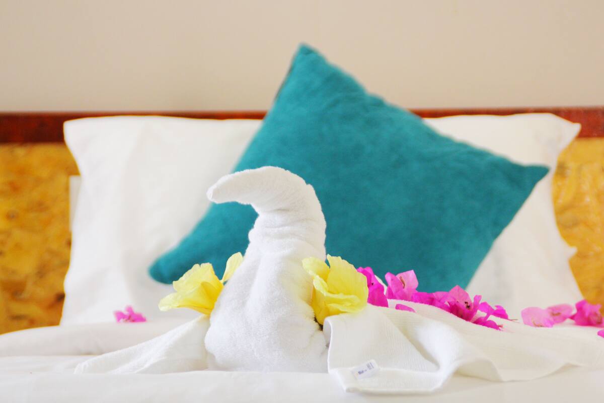 A bed with a white blanket, a teal pillow, and decorative flowers.
