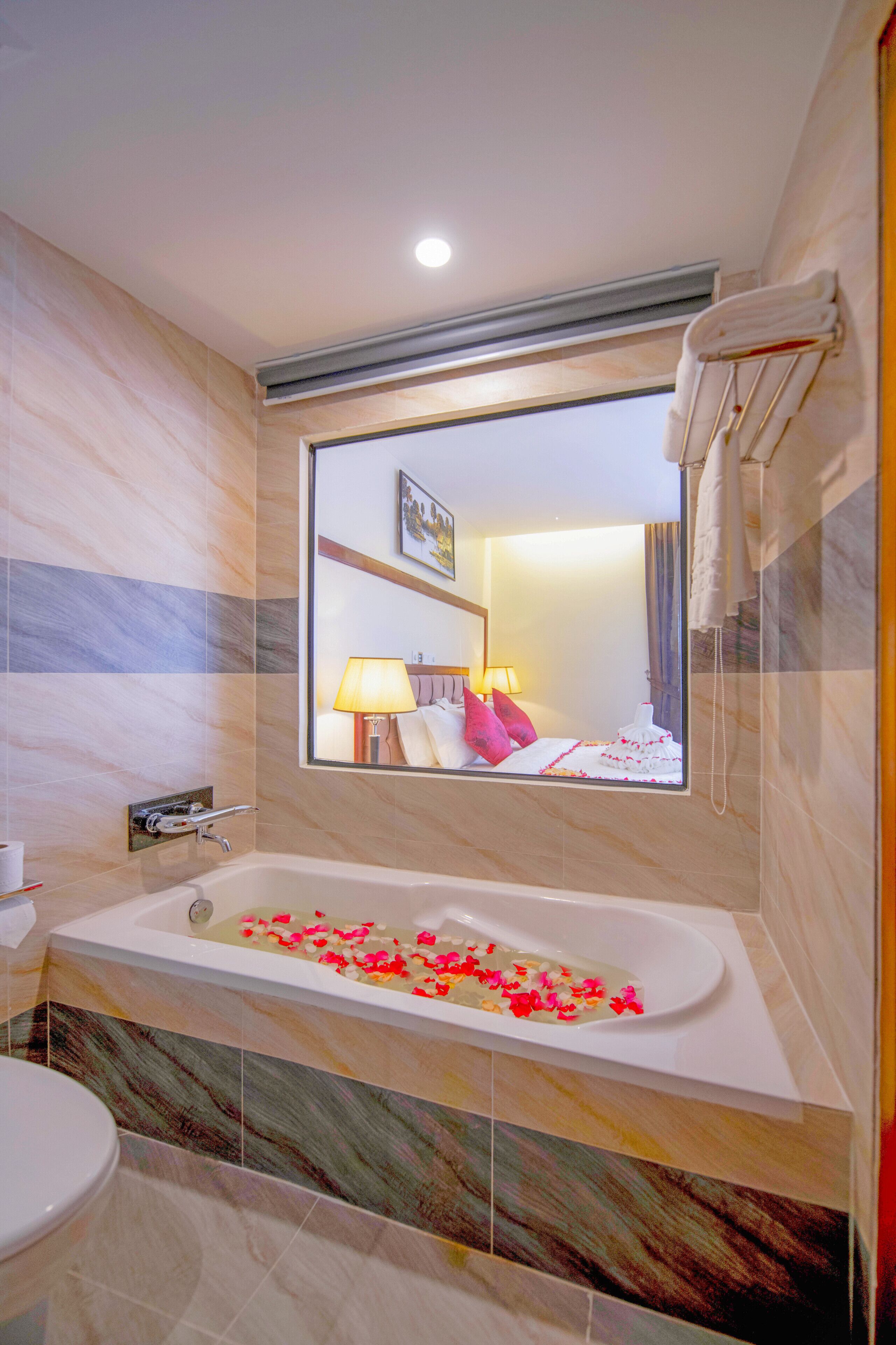 Ein Badezimmer mit einer Badewanne voller Rosenblätter, einer Toilette und einem Spiegel, der ein Bett mit Lampe und Kissen reflektiert.