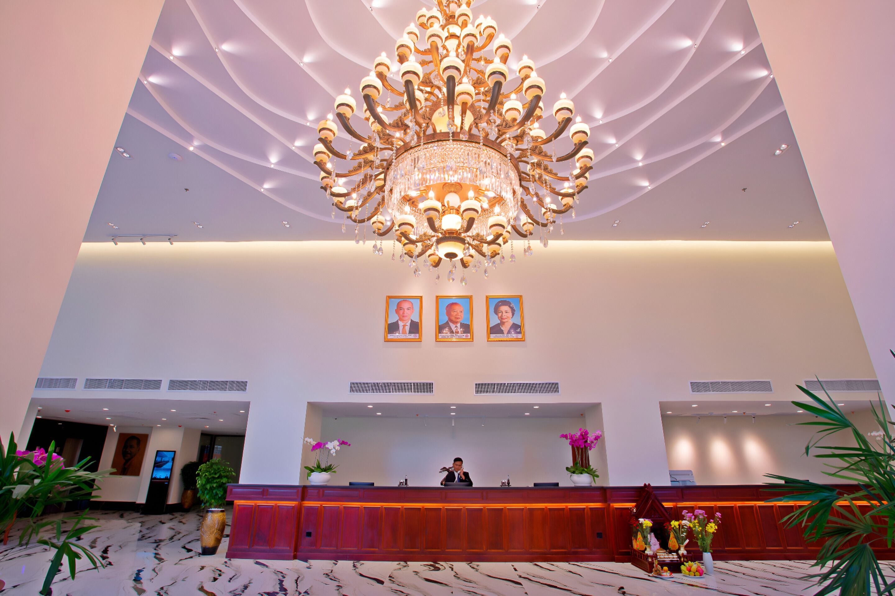 A hotel reception area with a large chandelier, a reception desk, and framed portraits above it.