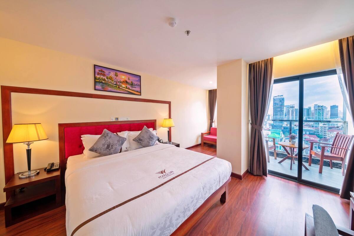 Habitación de hotel con una cama grande, vistas a la ciudad a través de la ventana, un cuadro en la pared y un balcón con sillas.